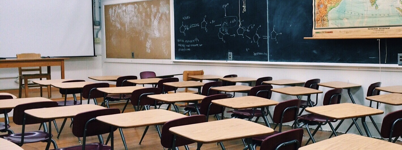clase con pupitres y sillas - al fondo una pizarra blanca detrás de la mesa y silla del profesor
